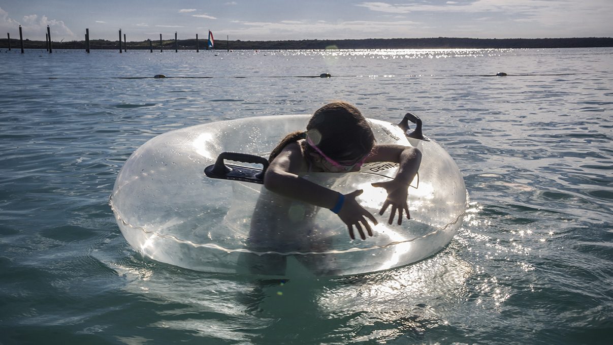 A child playing in a clear inflatable tube in a calm body of water on a sunny day.